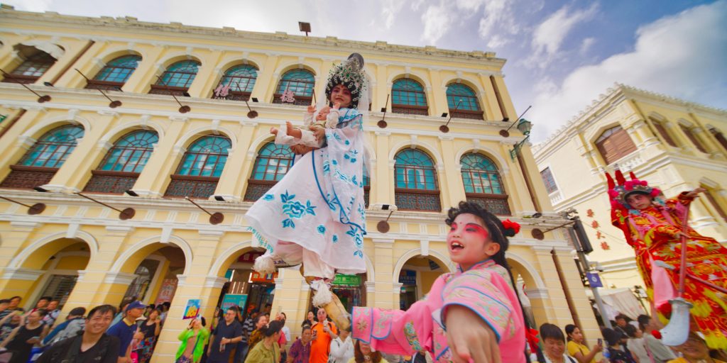 A little girl dressed as a deity holds a baby doll, a nod to Na Tcha as the protector of children